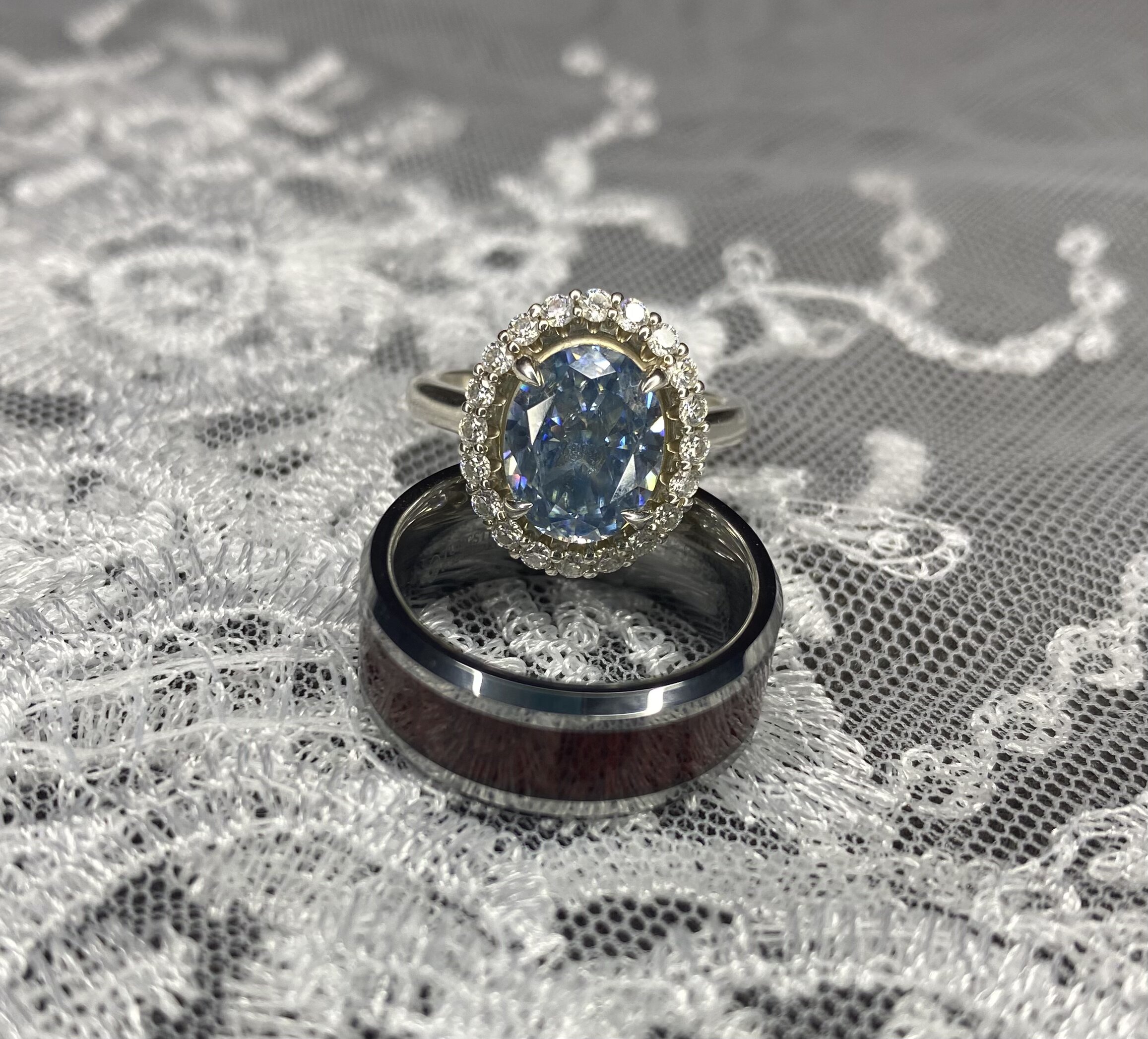 An ovan blue moissanite engagement ring standing on a wooden wedding band, on white lace